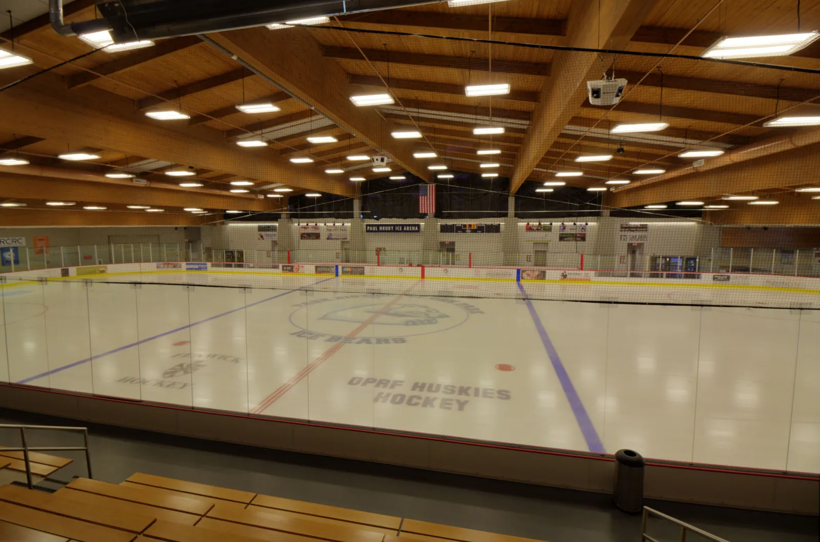 The empty rink from the stands, with "OPRF Huskies Hockey" painted on the ice under a wooden roof structure lit by rows of ceiling lights. — Paul Hruby Ice Arena