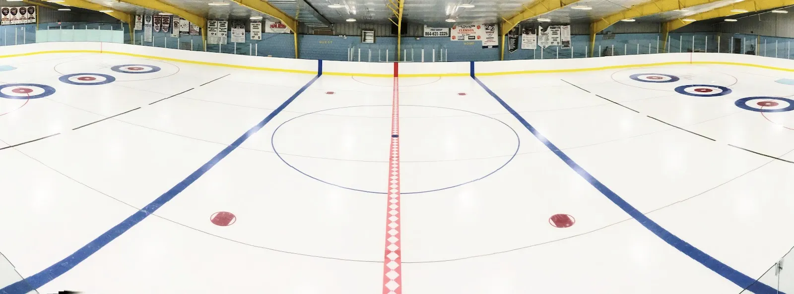La pista vacía vista desde detrás de las vallas del extremo, con la línea central roja, las líneas azules y los círculos de saque bajo un alto techo blanco con una franja de asientos en el lado opuesto. — Pavilion Recreation Complex