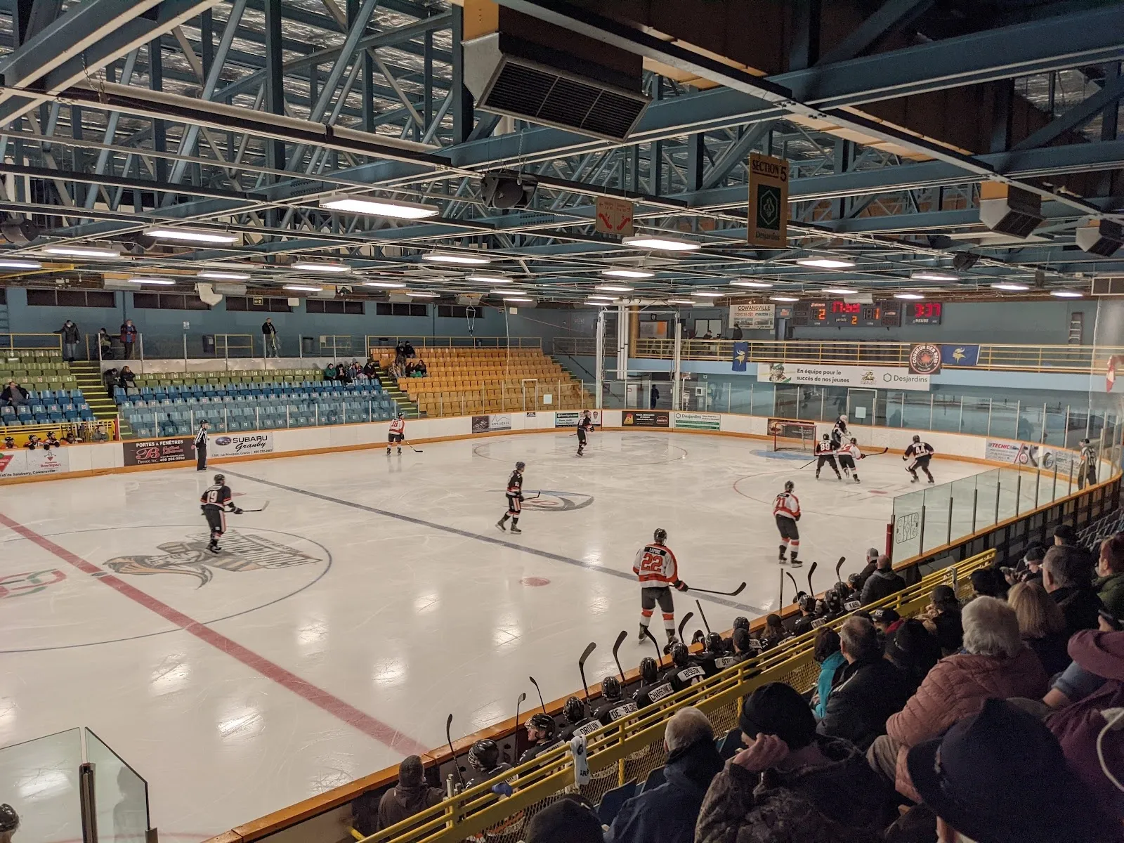 A game seen from the stands, players spread across the sheet with red-trimmed grandstands and a steel-trussed roof behind. — Pavillon des sports Roland-Désourdy