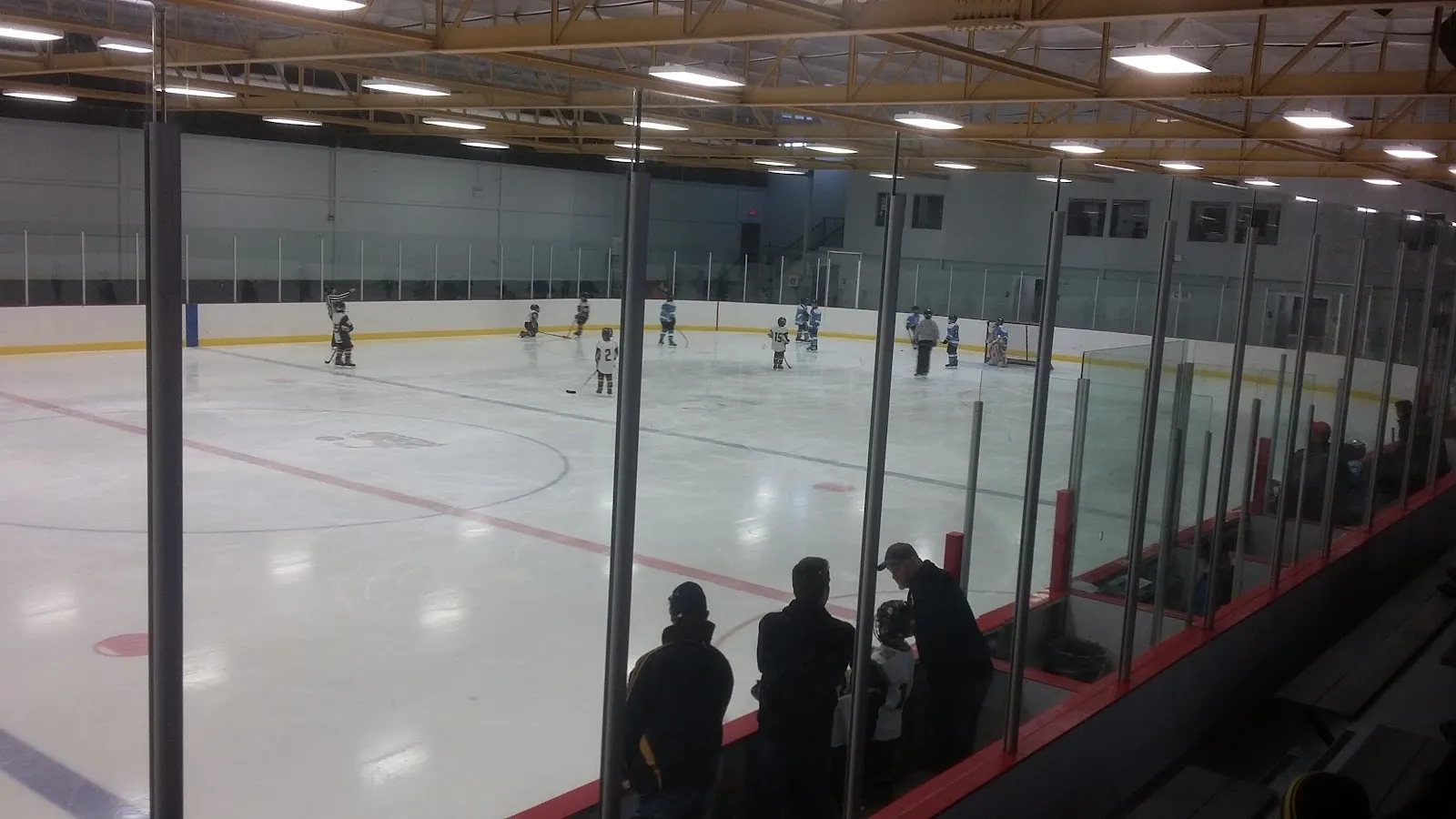 A youth hockey practice on the full ice sheet at the Pavillon sportif de Kénogami, seen from the stands through the glass — Pavillon sportif de Kénogami