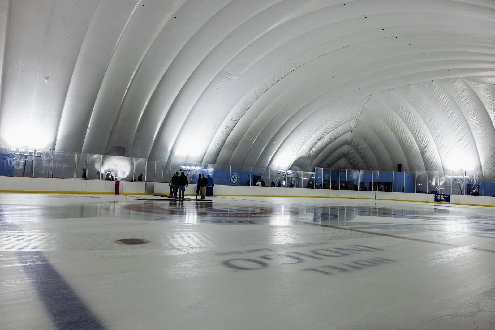 The long white inflatable dome arching over a wide sheet, the ice lit from above with a centre-ice logo and a few people at the far end. — Peconic Ice Rinks