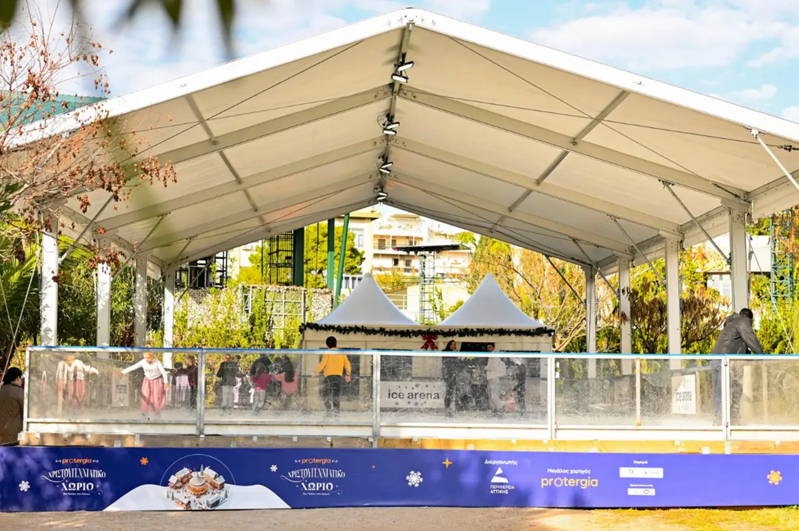 The covered ice rink at Pedion tou Areos under a white tent roof among the park trees, with small chalet roofs behind and a blue advertising barrier along the front. — Pedion tou Areos Ice Rink (Xmas Village Attica)