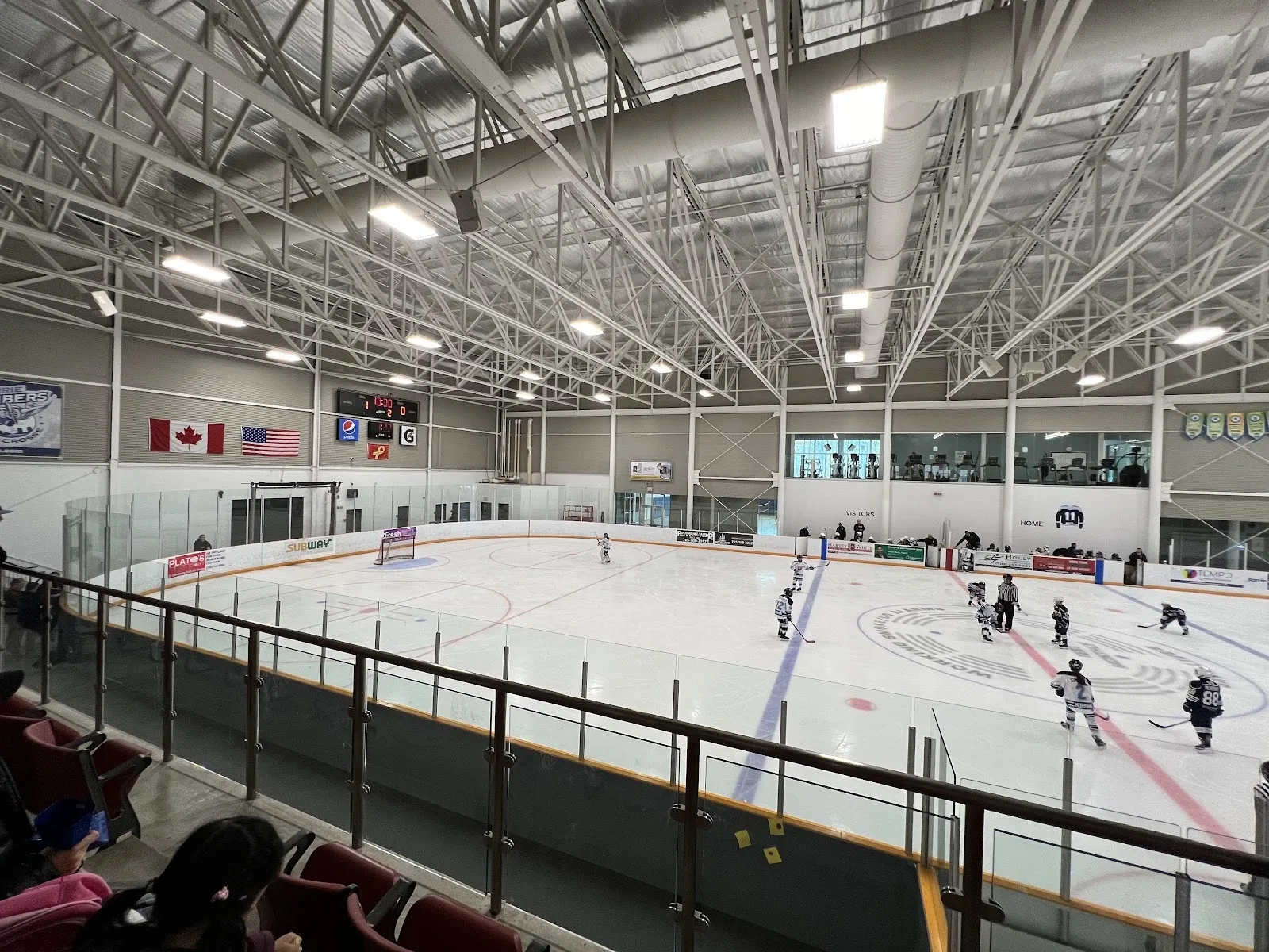 Una vista amplia de la pista desde un lado de las gradas rojas, con jugadores repartidos por el hielo blanco bajo un techo de armaduras de acero y banderas en la pared del fondo. — Peggy Hill Team Community Centre
