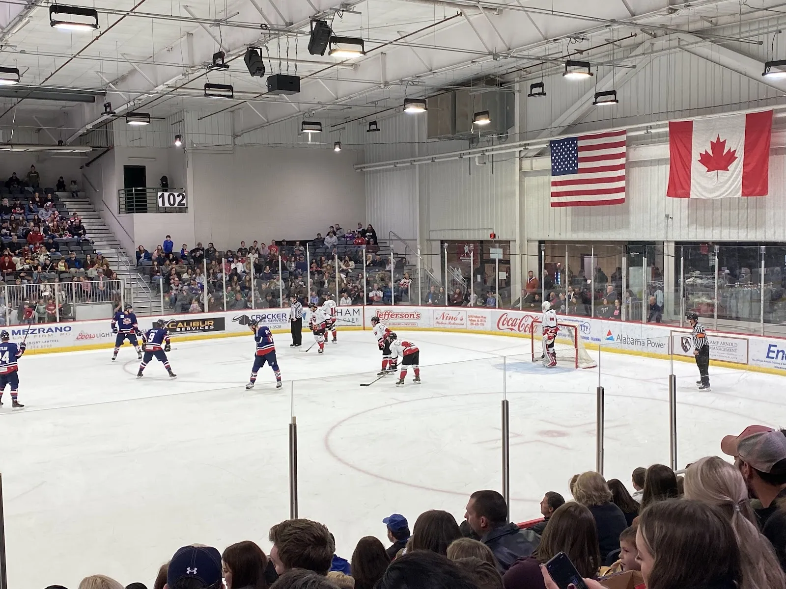 A game seen from the lower seats of the bowl, players spread across the sheet with American and Canadian flags on the far wall and a crowd filling the stands. — Pelham Civic Complex & Ice Arena