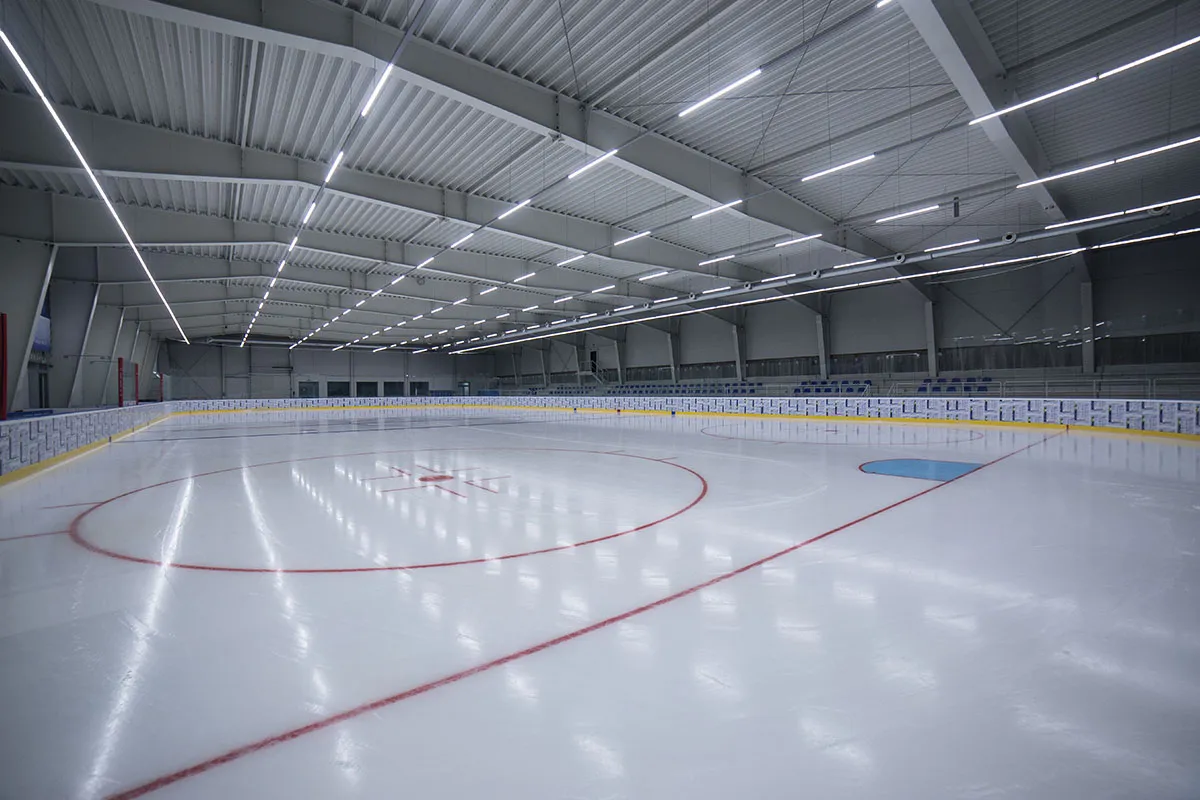 The empty ice sheet of the PEM arena seen from a corner, with the red centre line and face-off circle, a white and yellow board and a long ceiling of steel beams with strip lights. — PEM aréna Stará Ľubovňa