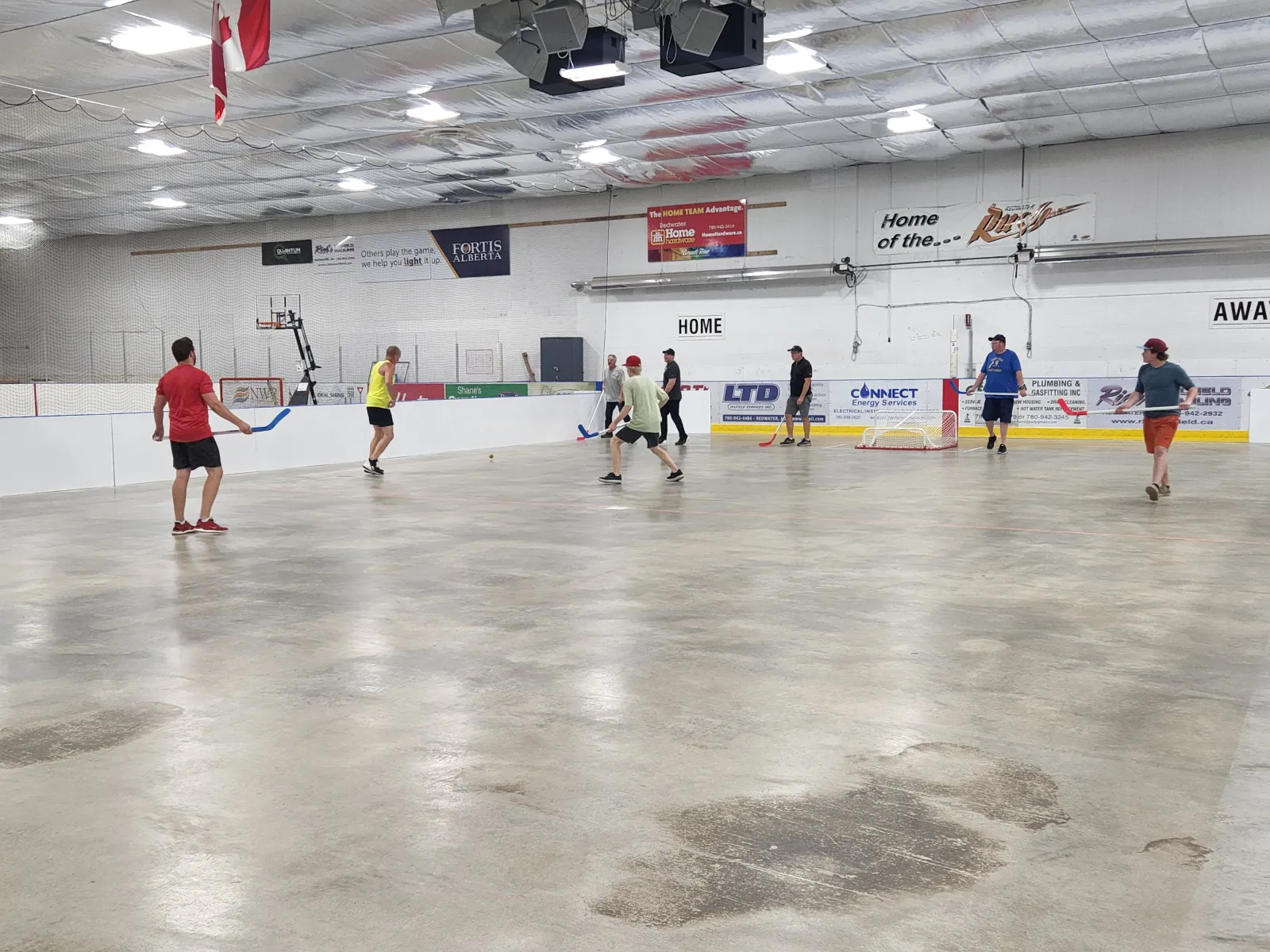 People playing with sticks across a wide, scuffed sheet, white boards with local sponsor signs and a Home sign along the far side. — Pembina Place