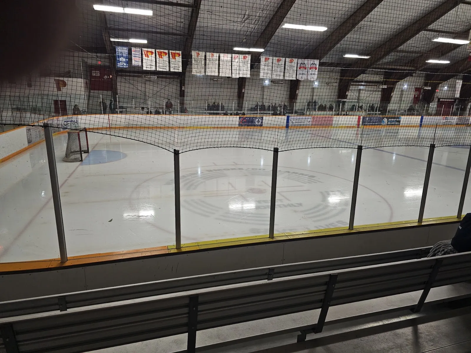 The ice seen through the end netting from the stands, with team banners along the roof. — Penetanguishene Memorial Community Centre