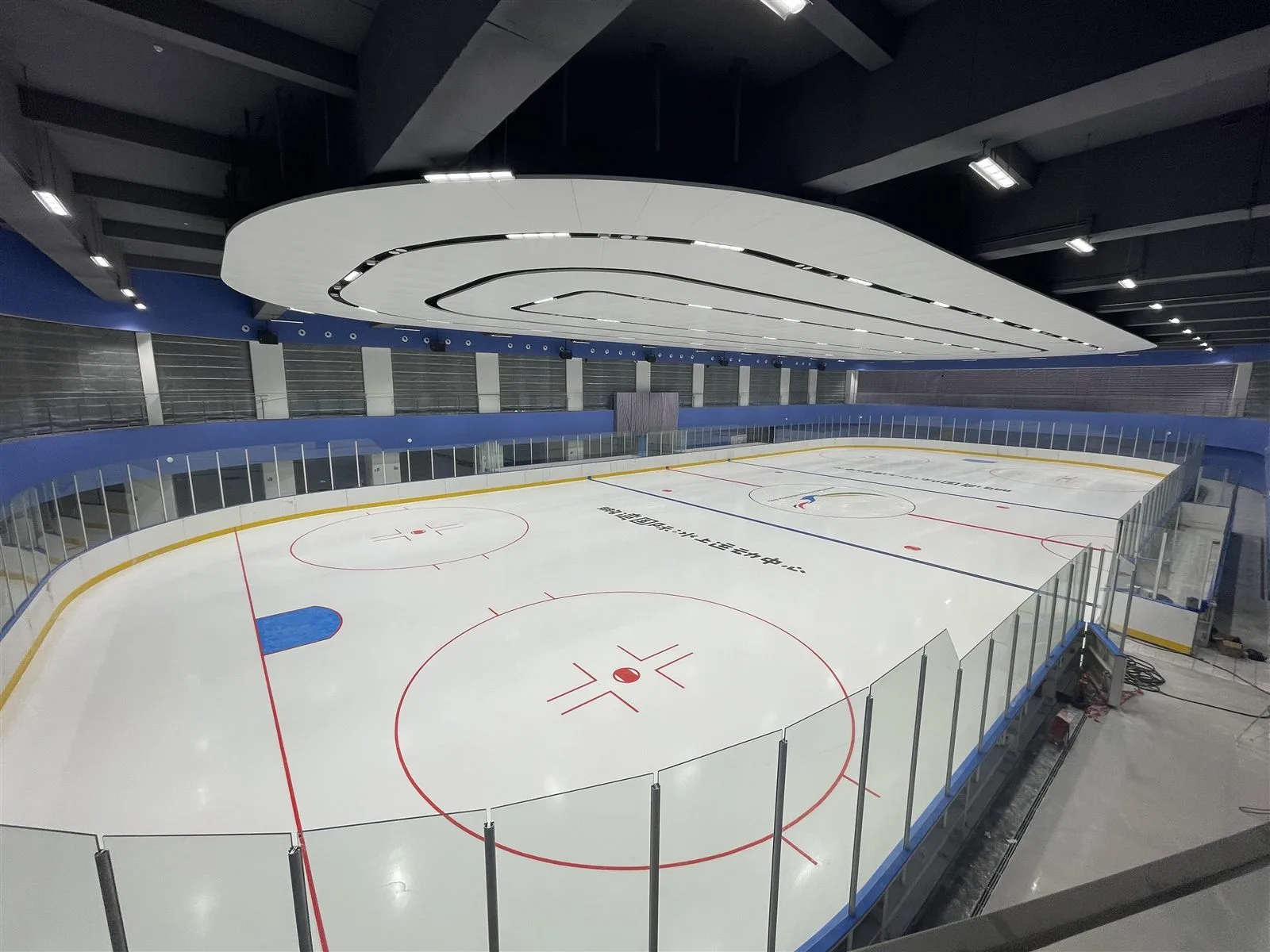 从角落望向空无一人的NHL标准冰场，玻璃界墙与冰面上方的椭圆形灯环 — Pengwan International Ice Rink