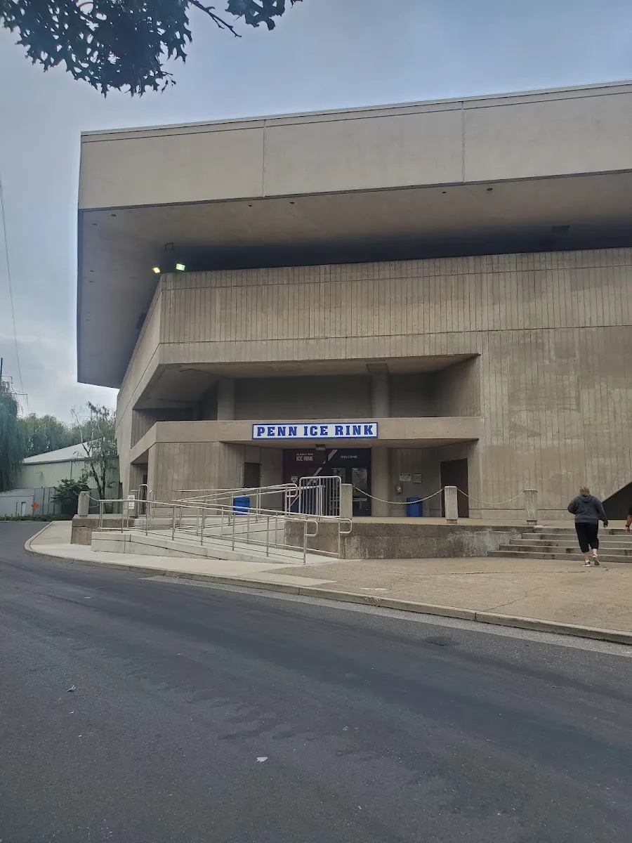 La arena de concreto aparente, con su nivel superior en voladizo y un letrero azul «PENN ICE RINK» sobre la entrada a nivel de calle. — Penn Ice Rink