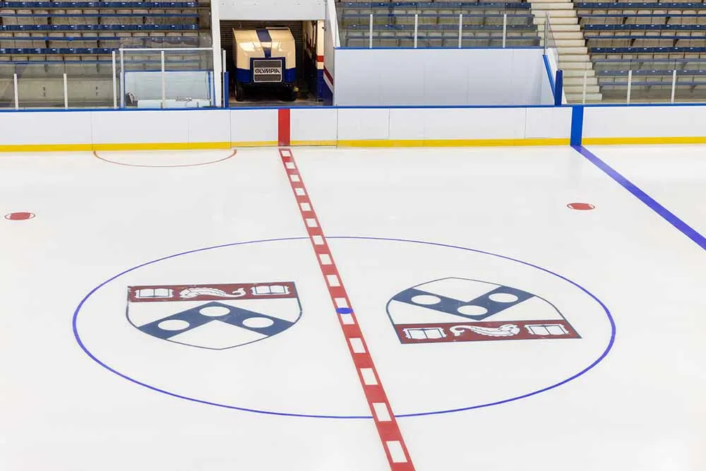 Hielo recién pulido al centro, con la línea central roja, el círculo azul y dos escudos de Penn pintados bajo la superficie, y asientos vacíos más allá de las bandas. — Penn Ice Rink