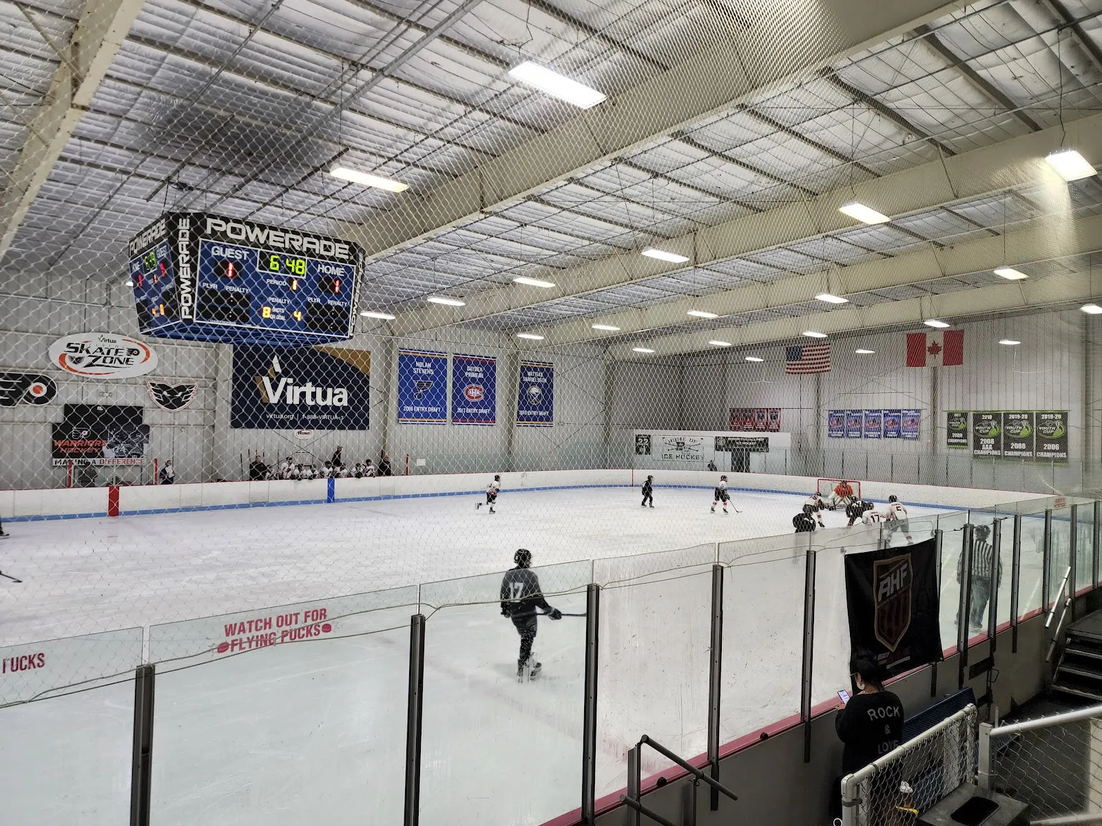 One of the two sheets seen from the stands through the netting, with the scoreboard overhead and banners along the far wall. — Pennsauken Skate Zone