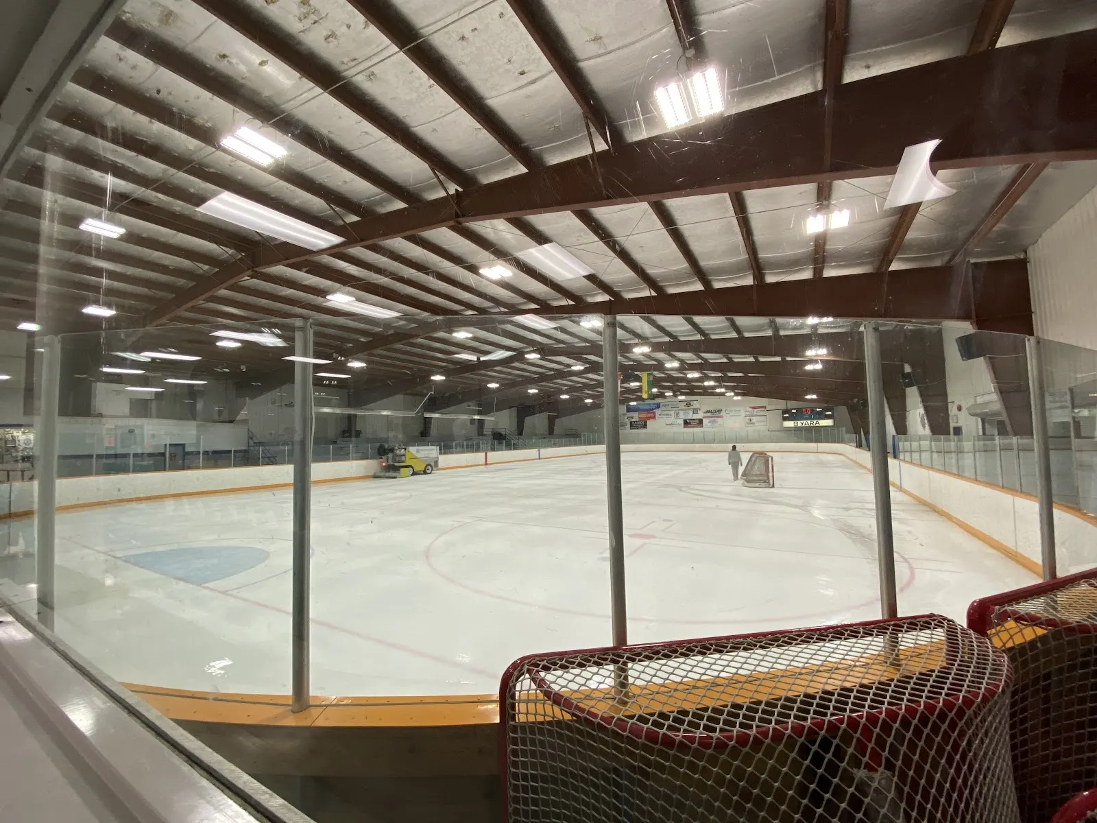 El hielo vacío visto a través del vidrio desde las gradas, con la Zamboni en el extremo del fondo y vigas oscuras de madera en el techo. — Pense Memorial Rink