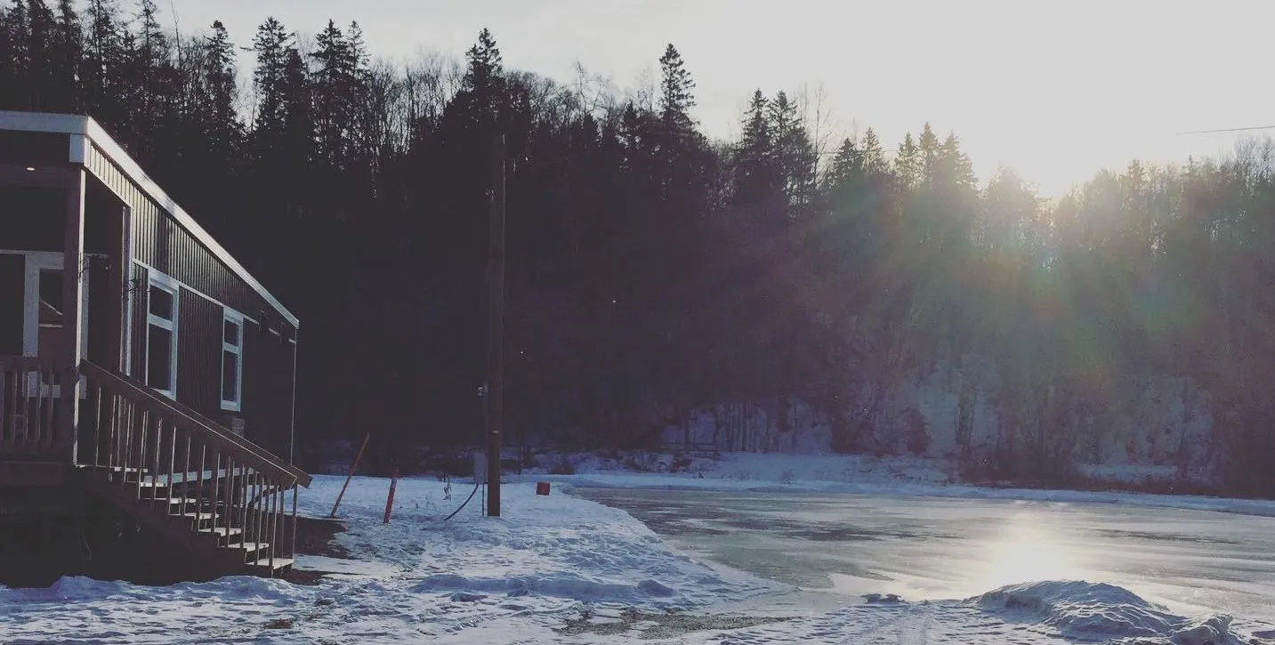 La pista exterior de Perth-Andover en una tarde de invierno: una superficie de hielo inundada bordeada de nieve al pie de una colina boscosa, con la caseta de calentamiento de la pista y sus escalones a la izquierda. — Perth-Andover Outdoor Rink