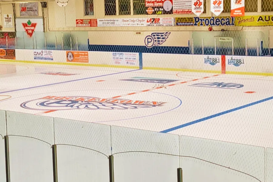 El hielo vacío desde detrás de las bardas, con el logotipo del centro, letreros de patrocinadores y mantas en la pared del fondo. — Perth & District Community Centre