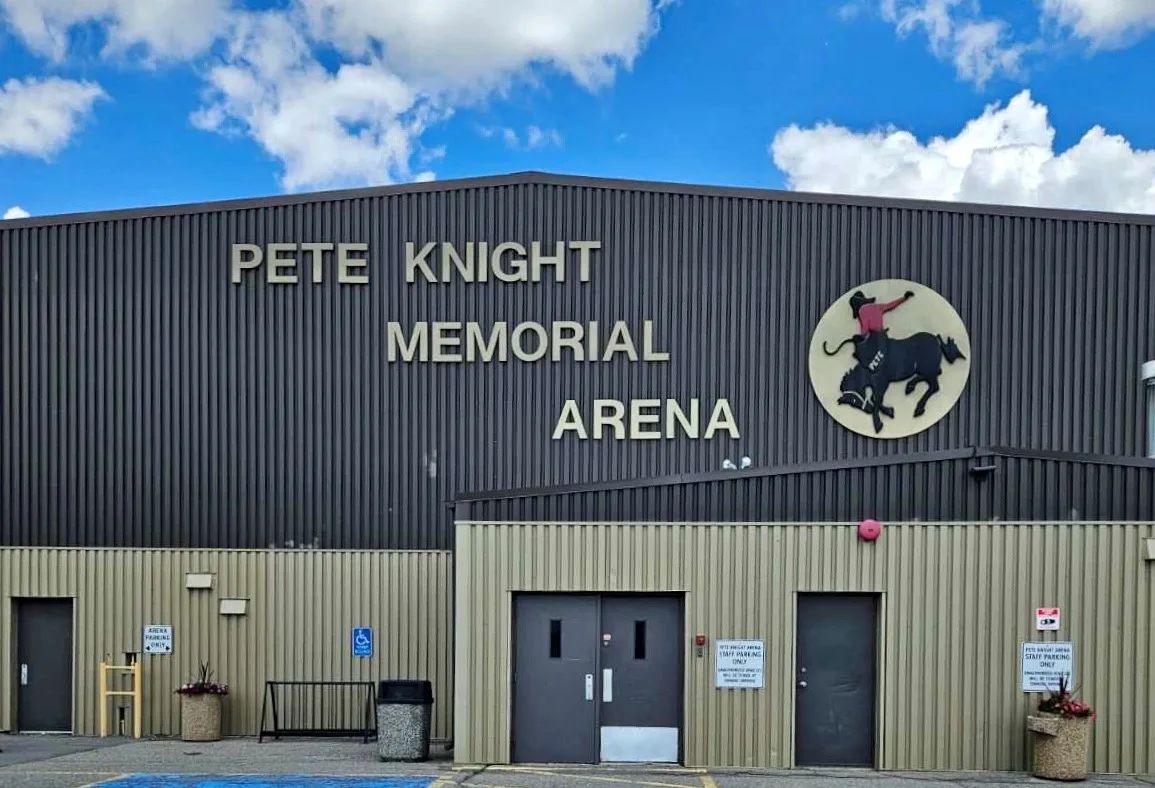 El muro frontal de metal acanalado oscuro con PETE KNIGHT MEMORIAL ARENA en letras grandes y un emblema de jinete de bronco, sobre puertas de entrada color arena y estacionamiento accesible. — Pete Knight Memorial Arena