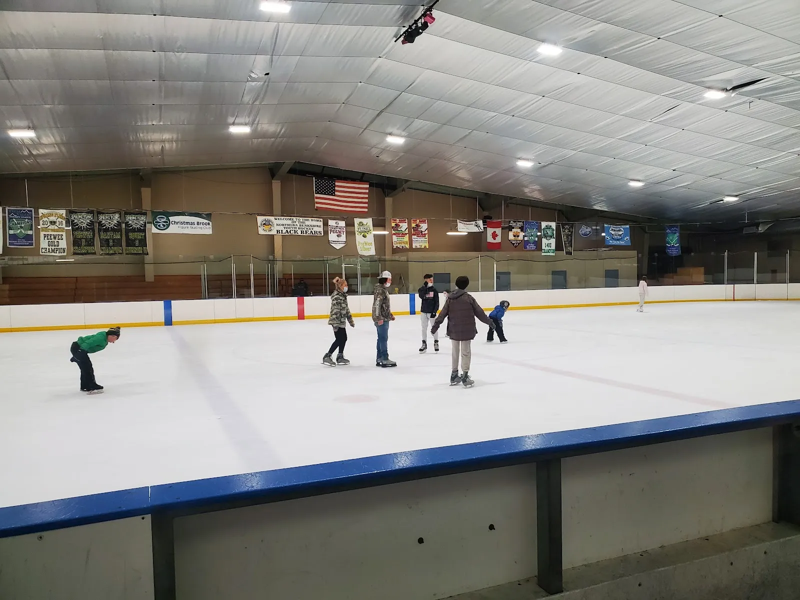 Toda la pista bajo un techo metálico a dos aguas, con mantas de campeonato y una bandera de Estados Unidos colgadas a lo largo de las vallas y unos cuantos patinadores en el hielo. — Peter W. Foote Vietnam Veterans Memorial Rink
