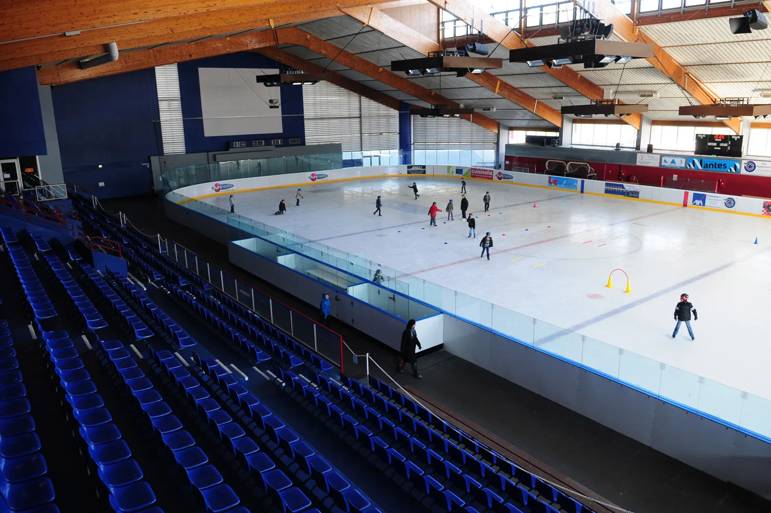The Olympic-size ice of the Petit Port rink seen from the stands, with a handful of skaters and play equipment on the sheet — Patinoire du Petit Port