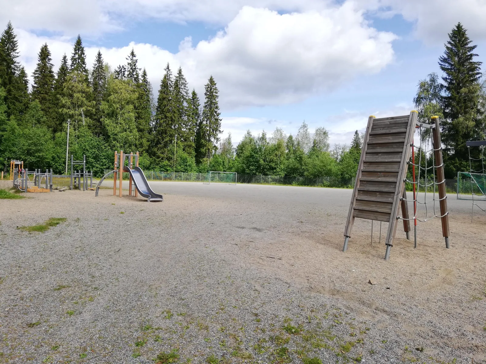 The gravel field at the Pihkainmäki school in Kuopio, which serves as a skating field in winter, seen in summer with a climbing frame and slide beside it and woods behind. — Pihkainmäen koulun luistelukenttä