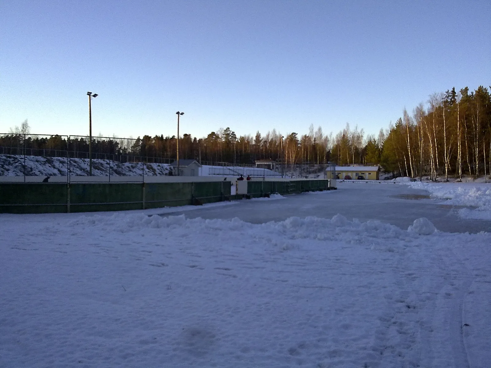 The cleared natural-ice field at the Pihlava sports ground in the snow, with the boarded hockey rink and floodlight poles behind and a birch and pine wood beyond. — Pihlavan luistelukenttä