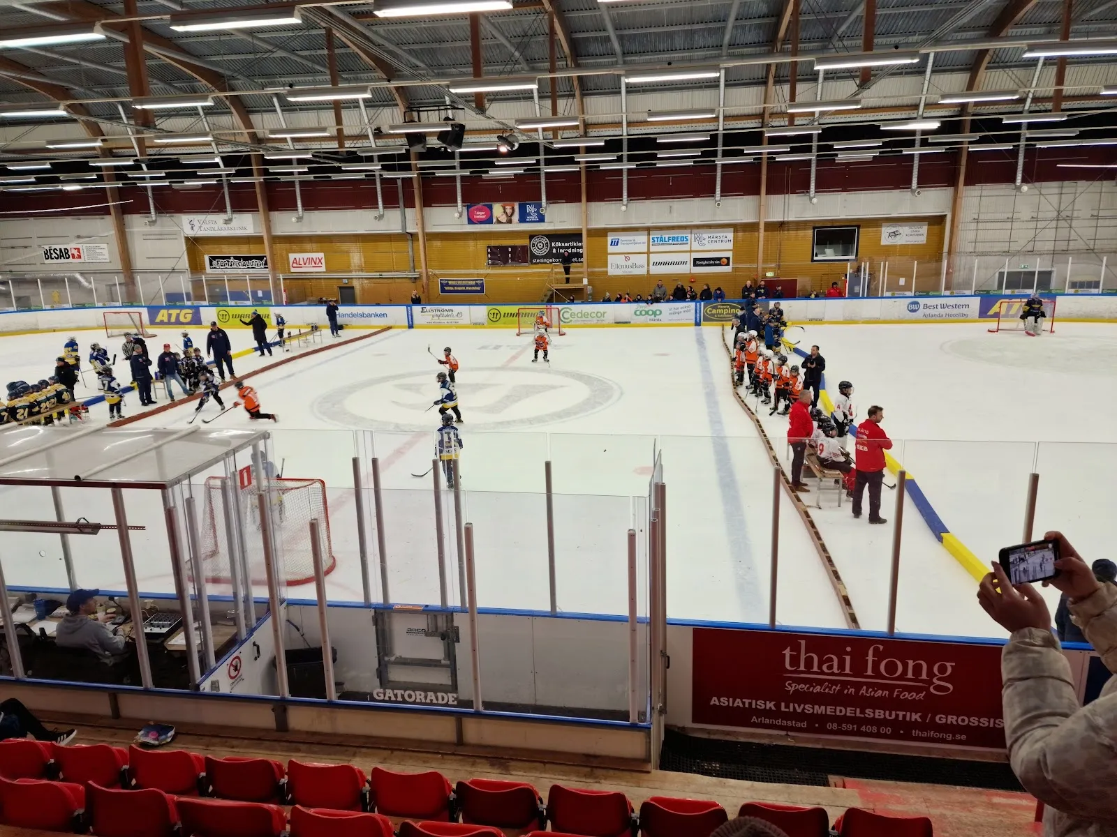 En ungdomsmatch på isen i Pinbackshallen i Märsta, sedd från sargen över isytan med målen och sponsortavlorna. – Pinbackshallen (Märsta)