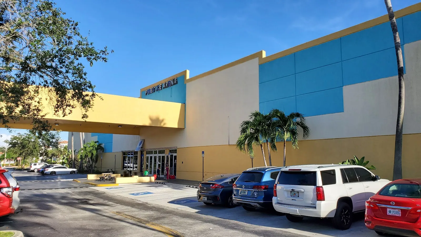 Exterior del edificio con paneles azules y color arena y un letrero azul en relieve de "PINES ICE ARENA" sobre la marquesina de la entrada, con el estacionamiento y palmeras al frente. — Pines Ice Arena (Pembroke Pines)