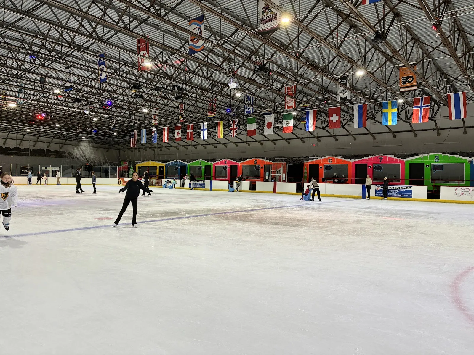 Toda la pista durante el patinaje público, con varios patinadores repartidos por el hielo, coloridos palcos de renta o fiestas numerados del 1 al 8 a lo largo de las vallas y banderas nacionales colgadas de las armaduras del techo. — Pines Ice Arena (Pembroke Pines)