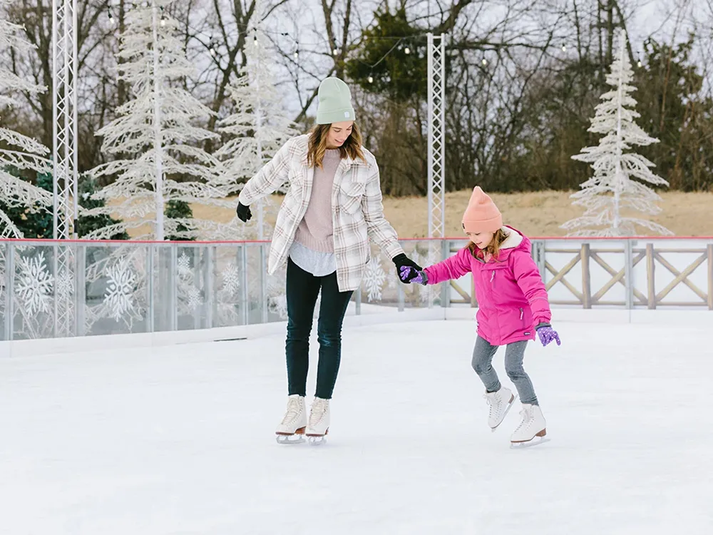 Una mujer y un niño patinan de la mano sobre el hielo blanco, con el cristal y las vallas de madera de la pista y árboles decorativos escarchados detrás de ellos. — Pinetop Ice Rink at Gaylord Opryland