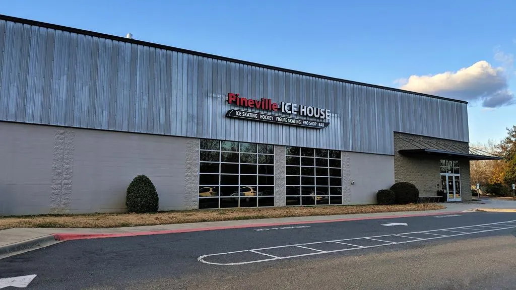 El edificio con revestimiento metálico de Pineville Ice House visto desde el estacionamiento, con un letrero rojo y blanco de 'Pineville ICE HOUSE' que enumera patinaje sobre hielo, hockey, patinaje artístico, pro shop y bar sobre grandes ventanales, y la entrada bajo un toldo a la derecha. — Pineville Ice House