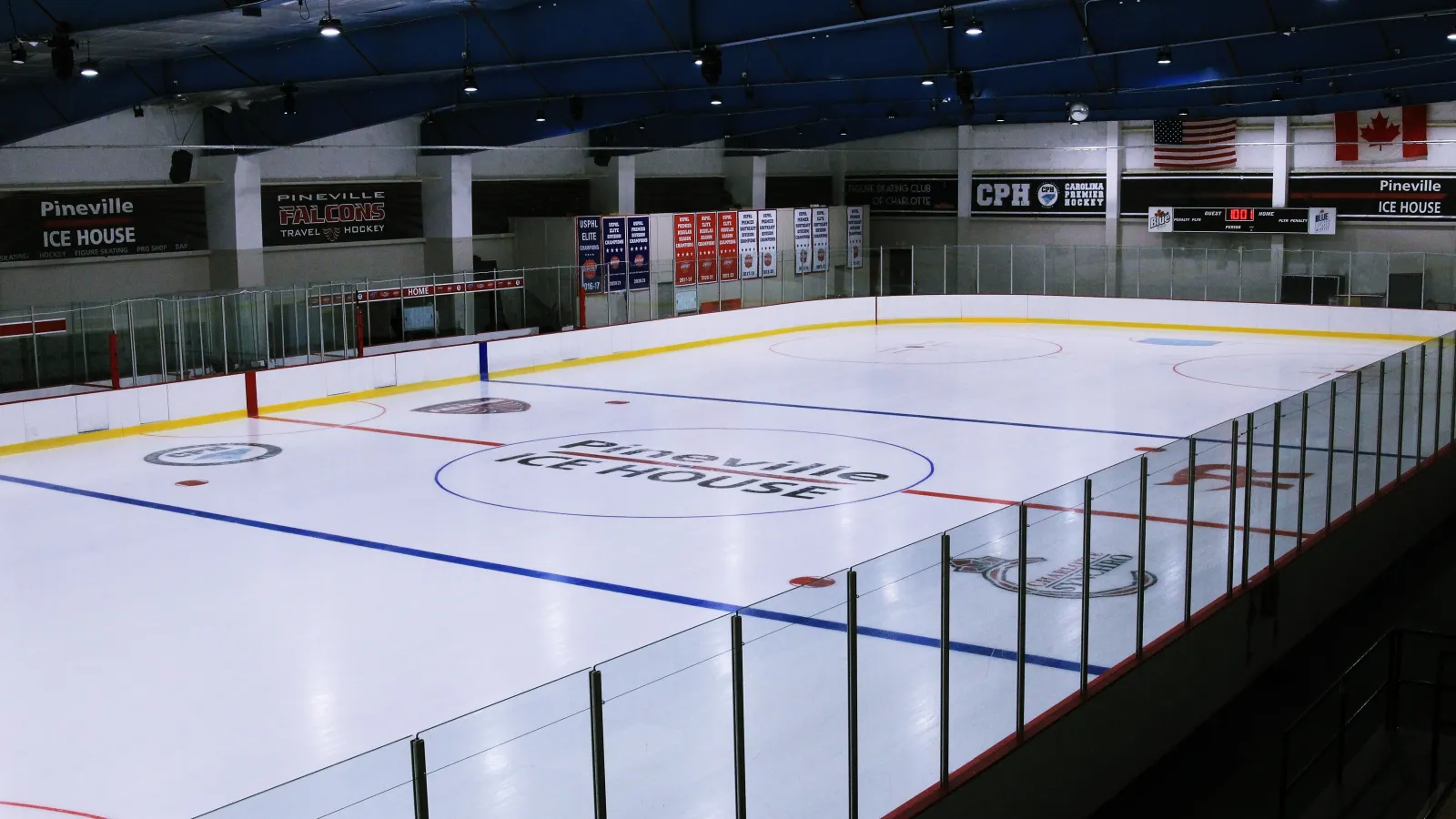 Pista vacía de Pineville Ice House vista desde una esquina elevada, con un logotipo de 'Pineville ICE HOUSE' en el centro de la pista y mantas junto a la pista de Pineville Falcons Travel Hockey, Figure Skating Club of Charlotte y Carolina Premier Hockey. — Pineville Ice House