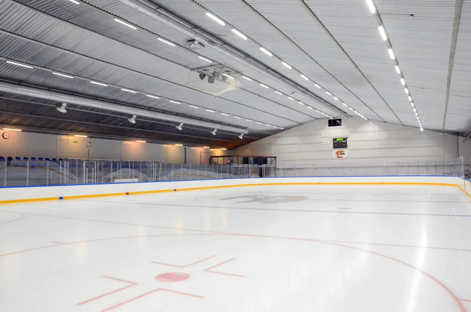 The empty ice of Pirkkalan jäähalli seen from the boards with the blue-and-red lines and face-off circle in the foreground and the grey hall wall and ceiling beyond — Pirkkalan jäähalli