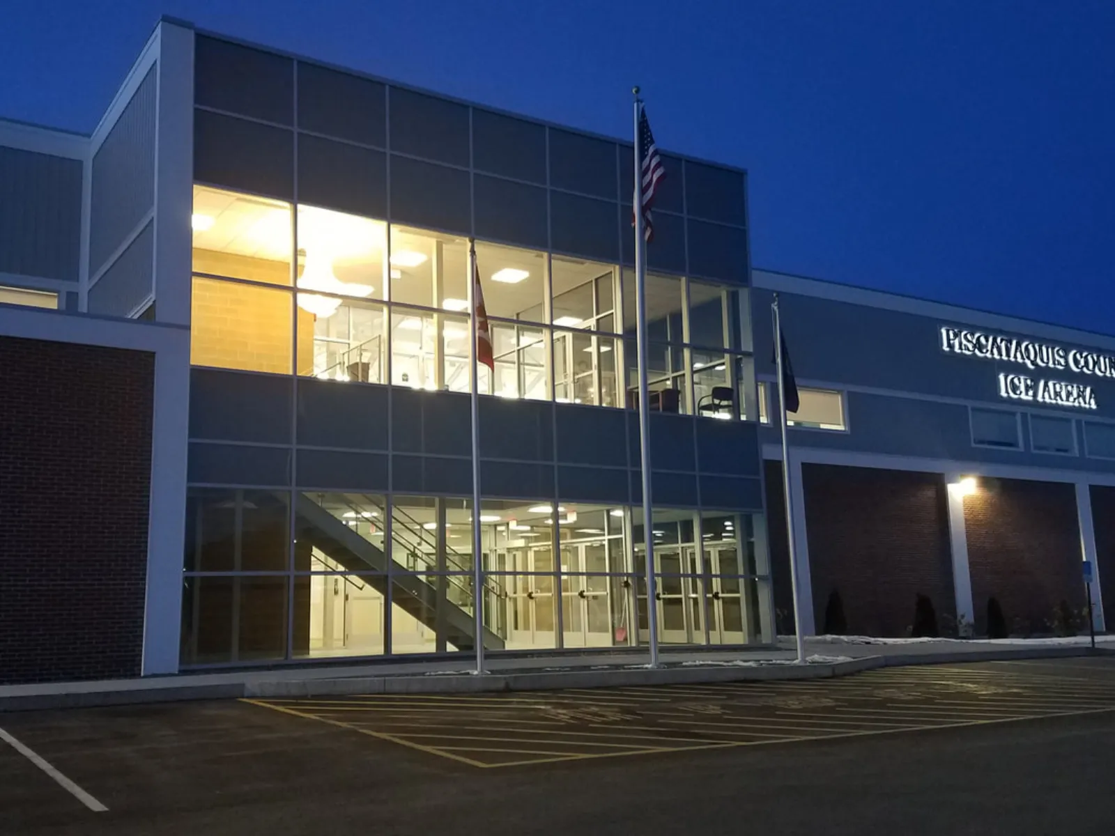 Dusk exterior of the two-story glass-and-metal front entrance, with illuminated "PISCATAQUIS COUNTY ICE ARENA" wall lettering, flagpoles, and the parking lot in the foreground. — Piscataquis County Ice Arena
