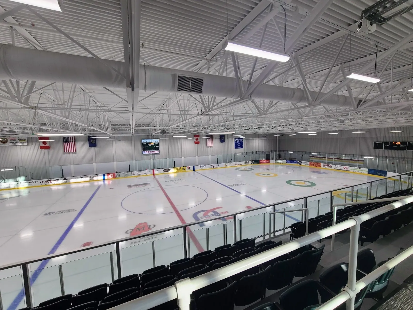 A wide view of the empty sheet from the stands, red and blue lines on white ice under a steel roof with a banner-lined end wall. — Piscataquis County Ice Arena