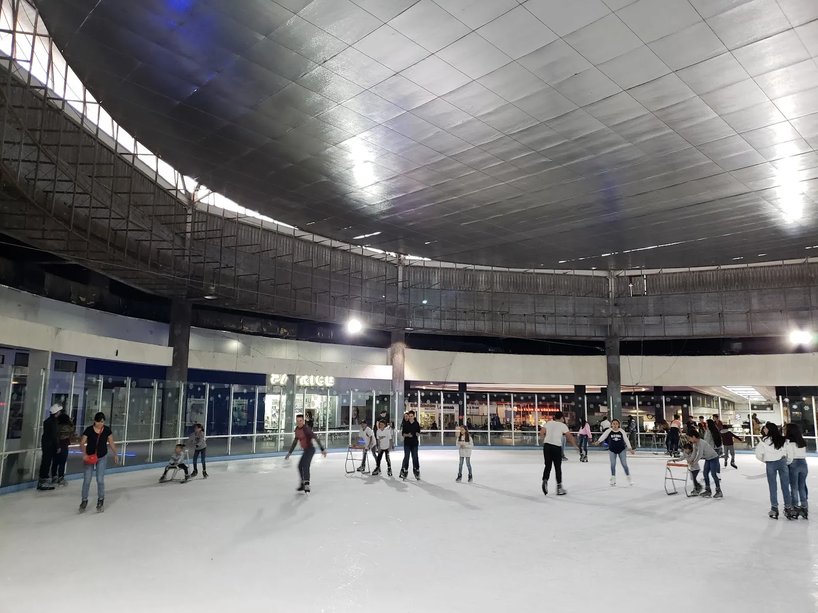 Ice rink under a curved metal roof, with skaters and skate aids across the sheet and shop fronts behind the boards. — Pista de Hielo Bugambilias