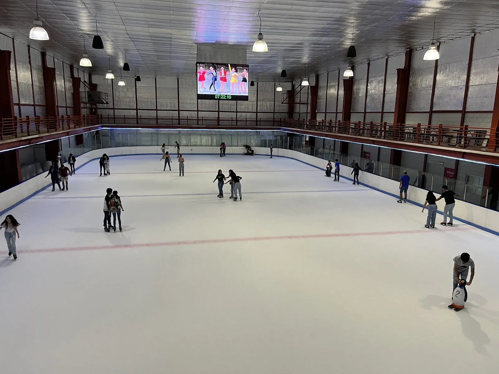 Ice sheet in a tall hall with a video screen above the far end and a balcony along the side walls, skaters spread across the ice. — Pista de Hielo Fundidora