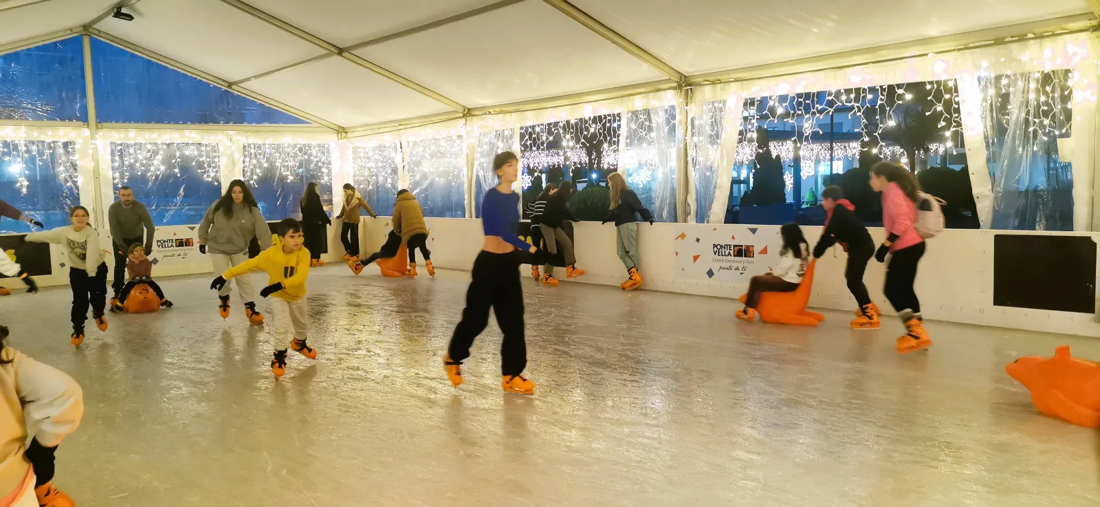 Skaters on the Ponte Vella ice rink under a white tent with lit curtains on the upper terrace, with children using orange seal walkers. — Pista de Hielo Ponte Vella