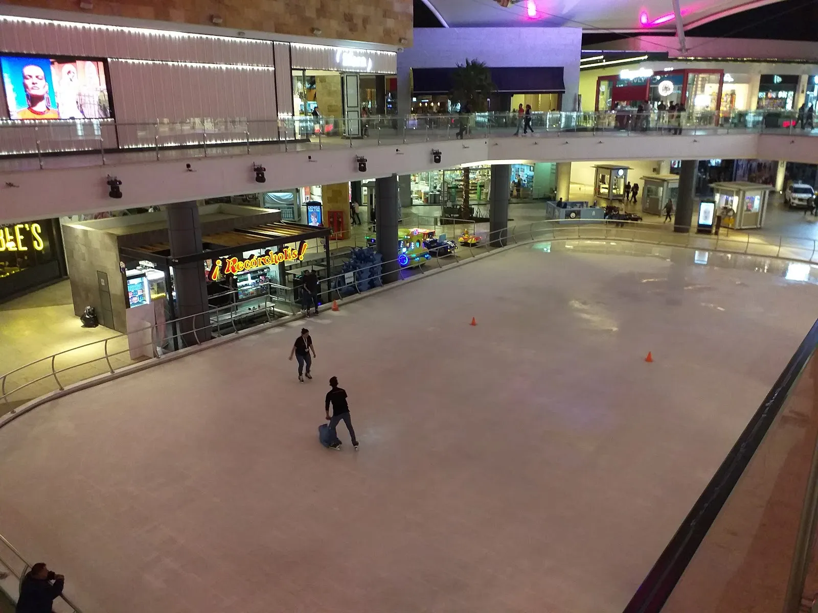 The mall rink at the Mundo E Recórcholis seen from the upper walkway, with two skaters on the wide ice and the Recórcholis kiosk and shops around it — Pista de Hielo ¡Recórcholis! Mundo E