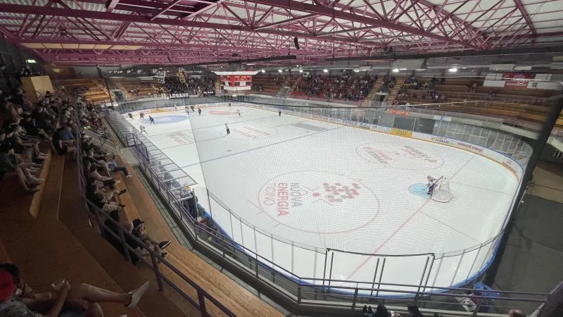 The covered BiascArena seen from the stands, with a game on the ice sheet and spectators along the boards. – Pista di Ghiaccio BiascArena