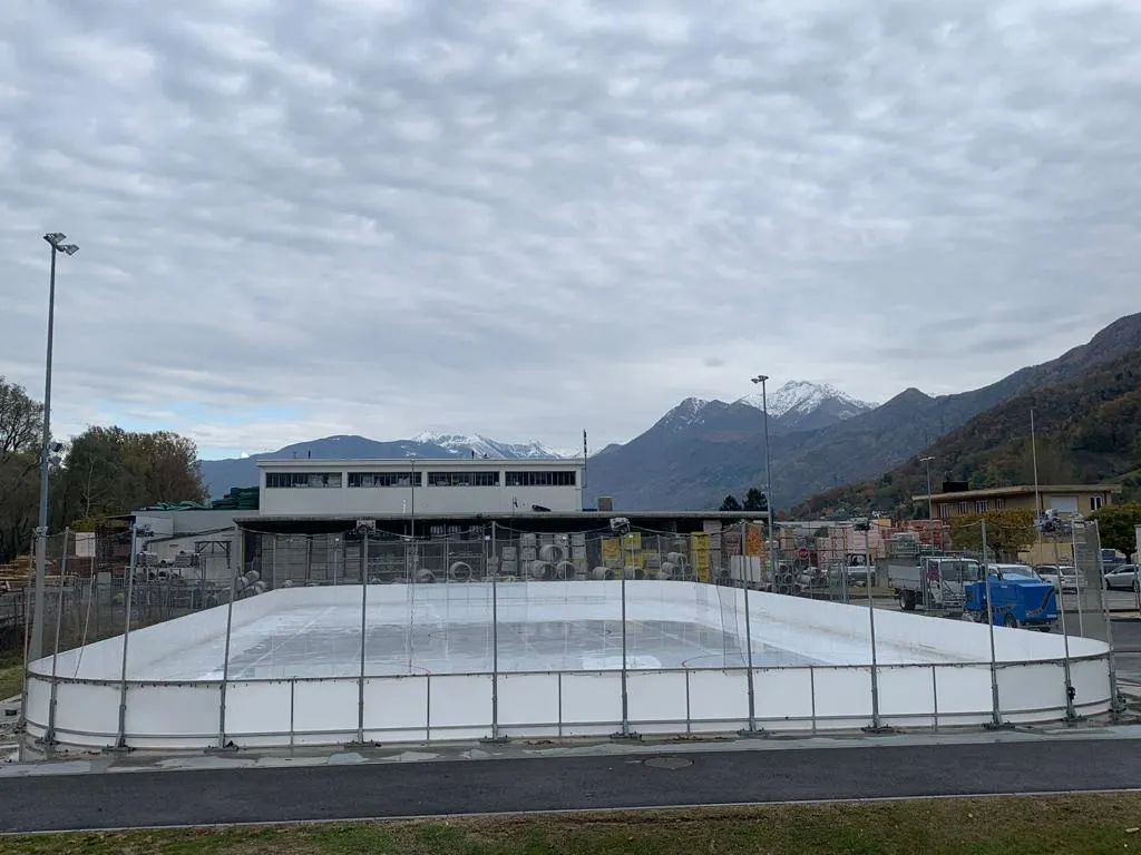 The boarded artificial ice rink at the Magadino sports centre, with the centre's buildings and the mountains of the Gambarogno behind. — Pista di ghiaccio Magadino