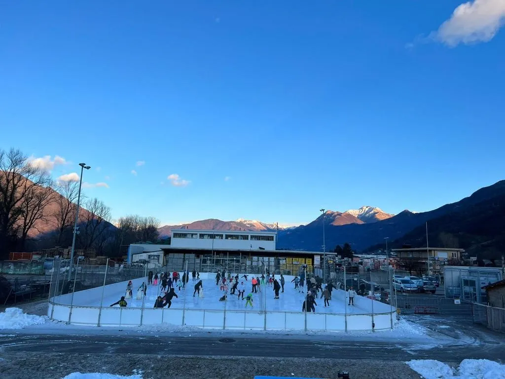 Skaters spread across the rink in the late afternoon, with the sports-centre building and snow-dusted peaks beyond the boards. — Pista di ghiaccio Magadino