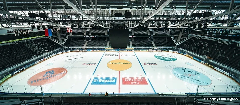 The empty Resega ice surface seen from above, with the centre-ice logos, the stands and the steel roof of the arena. — Pista del ghiaccio Resega