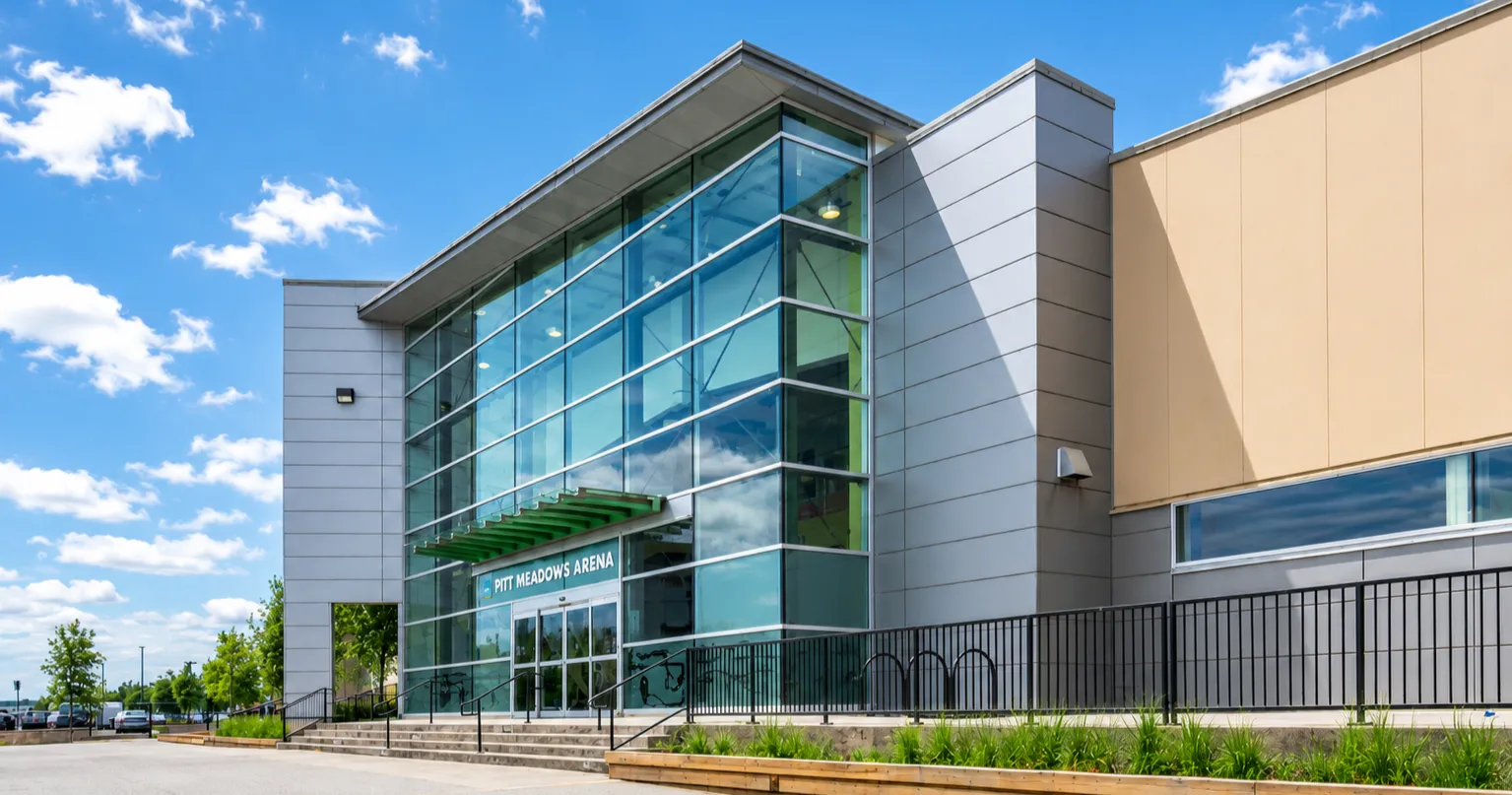 The glass-fronted entrance of the arena complex with the Pitt Meadows Arena sign, a landscaped bed and a railing along the walkway, under a blue sky. — Pitt Meadows Arena Complex