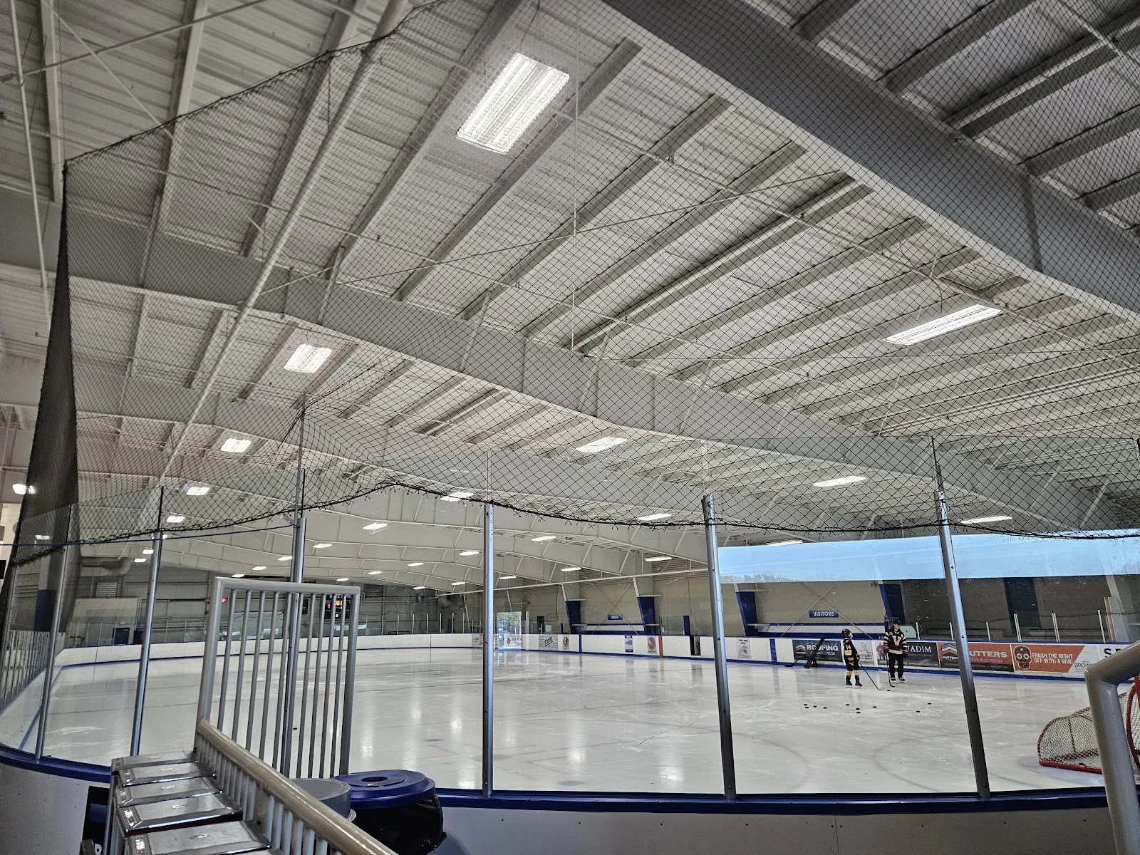The empty ice sheet at Pitt Meadows Arena Complex under a steel-truss roof, with the rink glass and a stair railing in the foreground. — Pitt Meadows Arena Complex