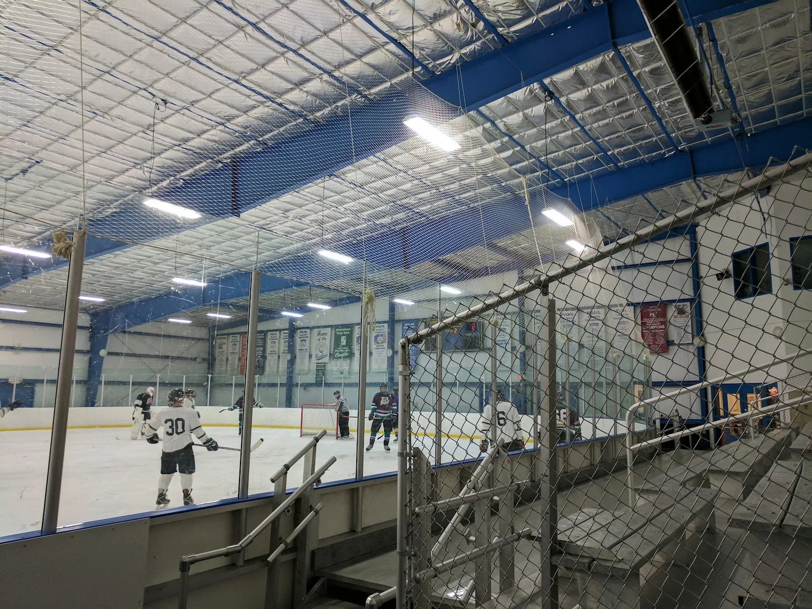 A game seen from the stands through a metal rail, players spread across the sheet under a blue-braced steel roof. — Pittsburgh Ice Arena