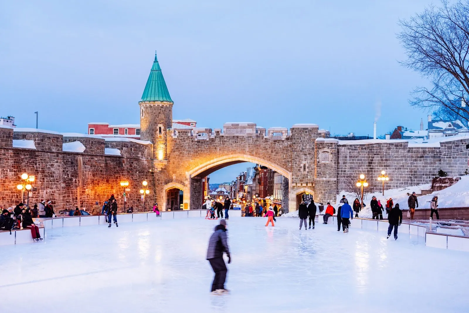 Des patineurs sur la patinoire de la place D'Youville en hiver au crépuscule, avec la porte Saint-Louis illuminée et l'ancien mur de la ville derrière la glace — Place D'Youville Outdoor Rink