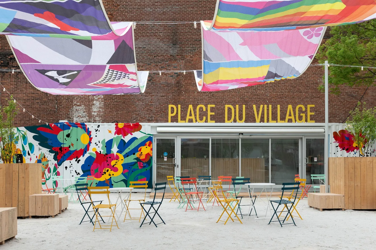L'enseigne de la place du Village au-dessus d'une murale et de tables de café sur la place recouverte de sable pendant la saison chaude, sans personne en vue — Place du Village Rink