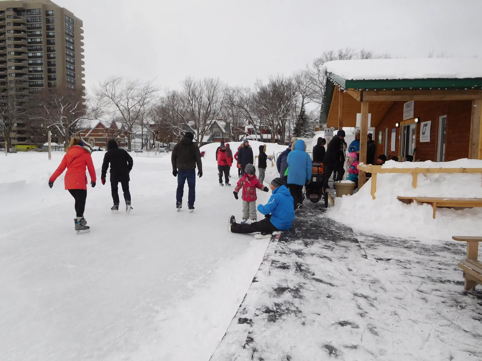 Skaters of all ages on the Plains of Abraham skating oval in winter, with the wooden chalet and apartment towers behind — Plains of Abraham Skating Oval (Anneau de glace des plaines d'Abraham)