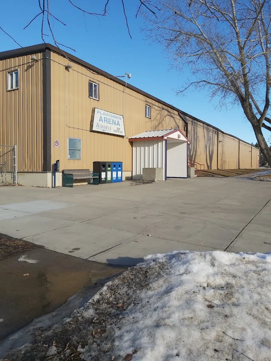 El lateral del Plainsmen Arena en un claro día de invierno: el largo edificio revestido de metal color arena con el rótulo Plainsmen Arena en 320 Centre Ave, la pequeña entrada cubierta, contenedores de reciclaje y un banco junto a la pared. — Plainsmen Arena