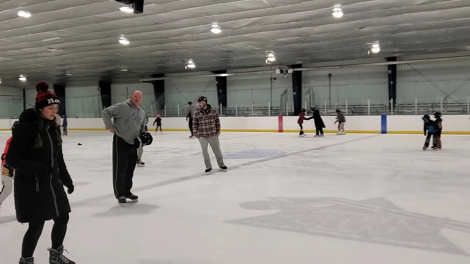 La superficie de hielo techada de la arena con patinadores por la pista y las vallas de la pared; la pista es el motivo principal. — Plainsmen Arena