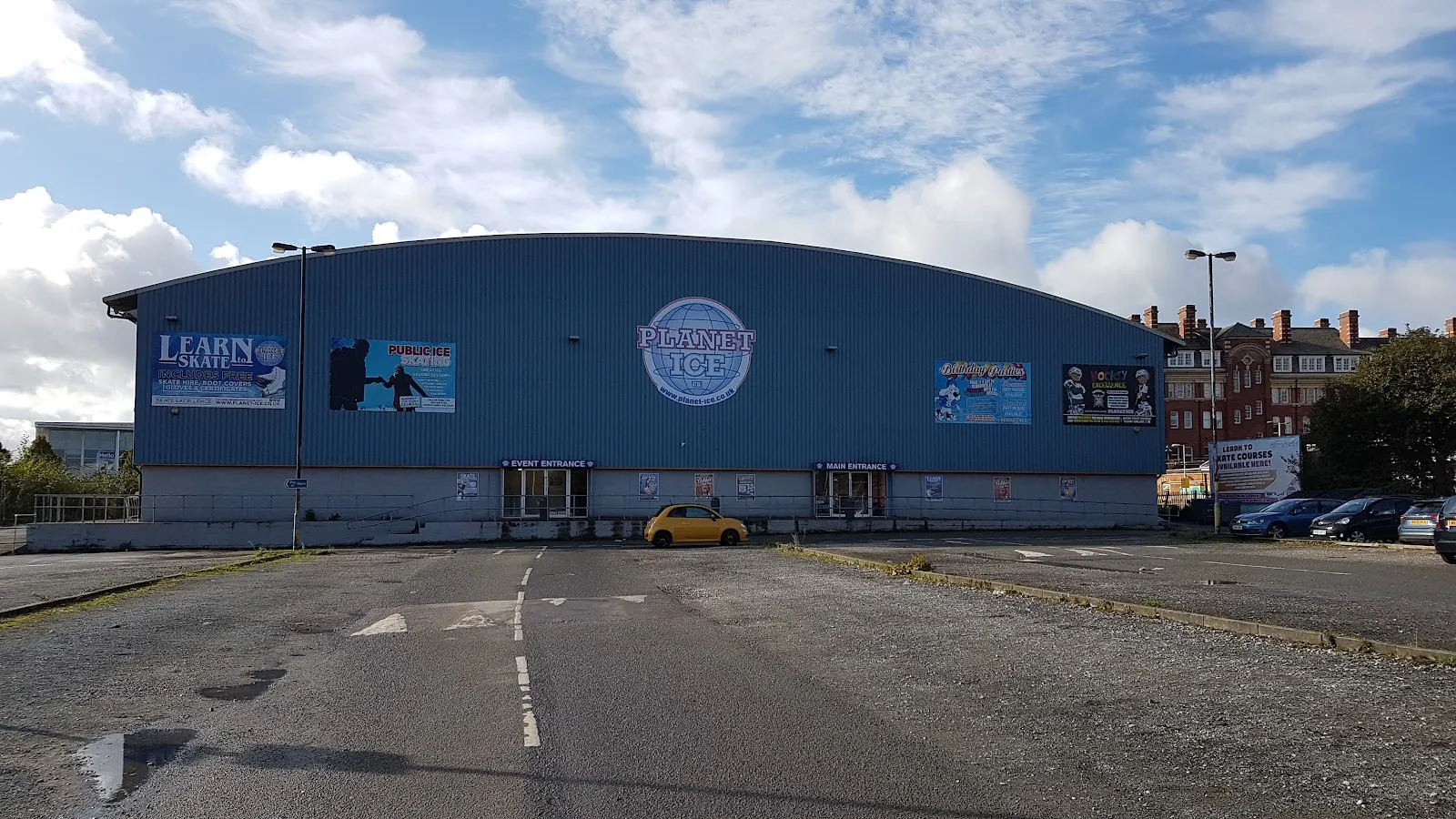 The long blue clad arch-roofed hall across its car park under a blue sky, with the Planet Ice globe and 'Learn to skate' signs on the front wall. — Planet Ice Altrincham