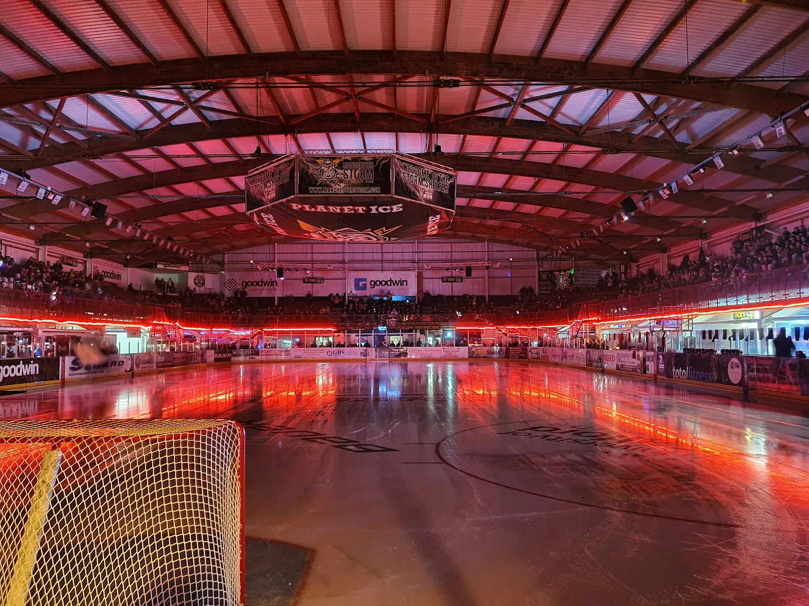 The empty ice sheet lit red and blue before a game, with a goal in the foreground, the scoreboard and the seating banks around the hall. — Planet Ice Altrincham